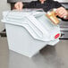 A man in a black coat using a scoop to put food into a white Rubbermaid ingredient container.