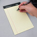 A hand using a pen to write on a yellow TOPS narrow ruled notepad.