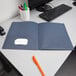 A dark blue Oxford 2-pocket folder on a desk with a computer and a white mug with pens in it.