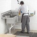 A man in a black shirt and grey pants cleaning a Regency stainless steel commercial corner sink on a counter in a professional kitchen.