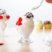 A person holding a spoon over a glass cup with ice cream and cherries.