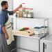 A man in an apron standing next to a Regency stainless steel table mounted overshelf in a professional kitchen.