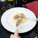 A person cutting a piece of food on a Tuxton Alaska plate.