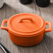 An orange porcelain oval cocotte with a handle on a table.