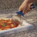 A hand with a blue polymer handle using a GI Metal stainless steel pizza cutter to cut a pizza on a table.