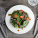 A white Bon Chef bone china salad plate with a spinach salad topped with tomatoes, onions, and feta.
