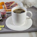 A white porcelain saucer with a cup of brown liquid on it and a plate of food.
