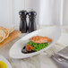 A slanted white porcelain bowl with a piece of salmon and peas on a table with a fork and knife.