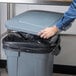 A person putting a black plastic bag into a Rubbermaid BRUTE trash can with a grey lid.