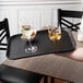 A hand holding a Cambro black non-skid serving tray with glasses of white wine and a bowl of ice cubes.