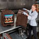 A woman in a white coat and gloves using a brown Cambro food container.