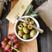 A bowl of green olives and grapes on a wooden cutting board with a Walco Luxor stainless steel cocktail fork.