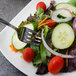 A Walco Pacific Rim stainless steel salad fork on a plate of salad with cucumbers, tomatoes, and lettuce.