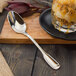 A Walco Luxor stainless steel dessert spoon on a cutting board next to a glass of dessert.