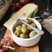 A bowl of green olives and grapes on a wooden cutting board with a Walco stainless steel cocktail fork.