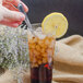 A hand holding a Walco stainless steel iced tea spoon over a glass of iced tea with a lemon slice.