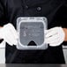 A person in white gloves holding a Carlisle clear plastic container with a hinged lid.