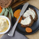 A Walco Bosa Nova stainless steel butter spreader with food and butter on a plate.