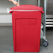 A woman opening a red Rubbermaid Slim Jim trash can with a plastic bag over it.