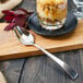 A Walco Freya stainless steel dessert spoon on a wooden cutting board next to a glass of food.