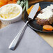 A Walco stainless steel butter spreader on a plate with butter, bread, and fruit.
