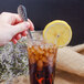 A hand holding a Walco stainless steel iced tea spoon over a glass of ice tea with a lemon slice.