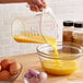 A person pouring orange liquid from a bowl of eggs into a clear plastic measuring cup.