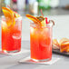 Two Acopa highball glasses filled with red liquid and fruit on a table in a cocktail bar.