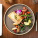 A Chef & Sommelier Geode gray stoneware dinner plate with salmon and vegetables on a table with a flower.