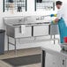 A man in a blue uniform cleaning a Regency 3 compartment stainless steel commercial sink.