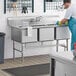 A man in blue gloves washing dishes in a Regency stainless steel 3 compartment sink.