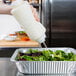 A hand pouring white sauce from a white FIFO squeeze bottle onto a salad in a foil tray.