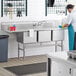 A woman in a blue uniform washing dishes in a Regency stainless steel 3 compartment sink.