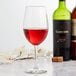 A Libbey wine glass filled with red wine on a table with bottles of wine.