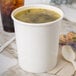 A white Choice paper cup filled with soup on a table with a spoon.