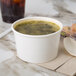 A white paper bowl of soup and a spoon on a marble surface.