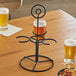 An Acopa black metal flight carrier with three glasses of beer and a plate of nuts on a table.