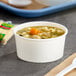 A bowl of soup in a white paper food cup with a spoon on a tray.