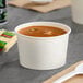 A white double poly-coated paper cup filled with soup on a table with a spoon.