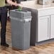 A woman standing next to a Lavex gray slim rectangular trash can with a plastic bag over it.