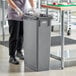 A woman in a chef's uniform standing next to a Lavex gray rectangular trash can.