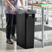 A woman in black gloves and an apron putting a black bag in a black Lavex Slim Rectangular Trash Can.