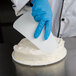 A person wearing blue gloves using a white Ateco triangular bowl scraper to frost a cake.