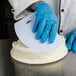 A person wearing blue gloves and using an Ateco large plastic bowl scraper to cut a white cake.