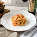 A white porcelain plate of pasta with shrimp next to a glass of wine.