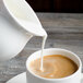 A white pitcher pouring milk into a cup of coffee.