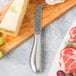 An American Metalcraft stainless steel cheese knife cutting a block of cheese on a wooden surface.