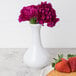 A white Arcoroc porcelain vase with purple flowers next to a plate of strawberries.