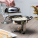 A person using a Libbey stainless steel chafing dish to pour cheese over food.