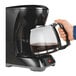 A hand holding a black Hamilton Beach coffee maker pouring coffee into a glass pot.
